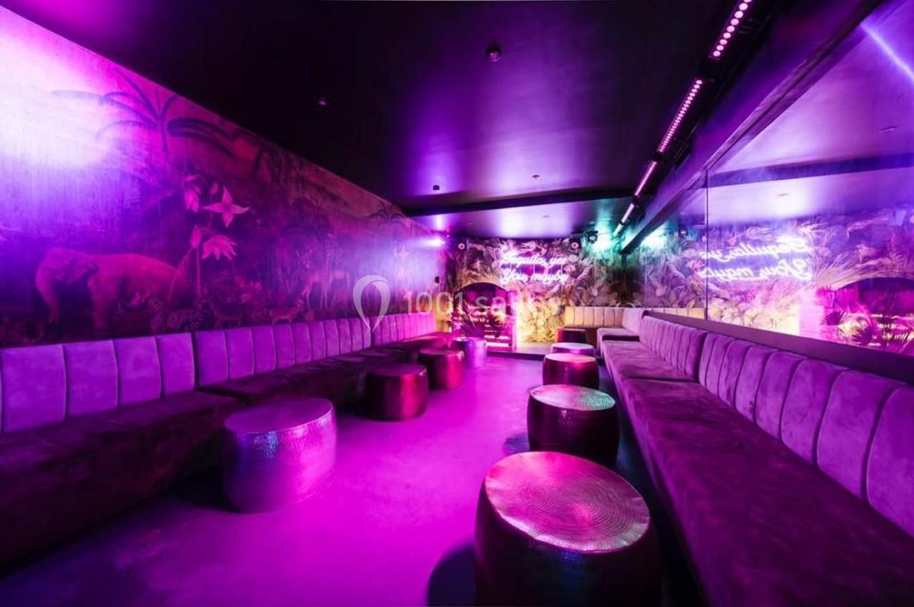 Salle lounge avec banquettes violettes, tables basses métalliques et éclairage néon rose et violet.