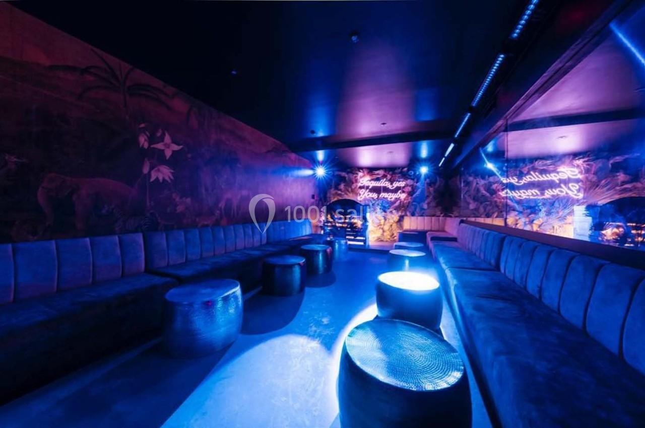 Salle lounge sombre avec banquettes, tables basses et éclairage bleu, décor mural tropical et néons lumineux.