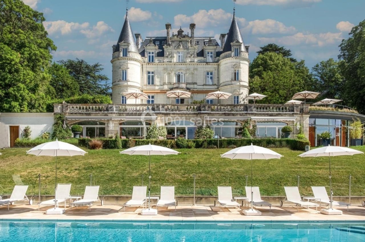 Façade d'un château entouré de verdure, avec une piscine et des transats alignés au premier plan.