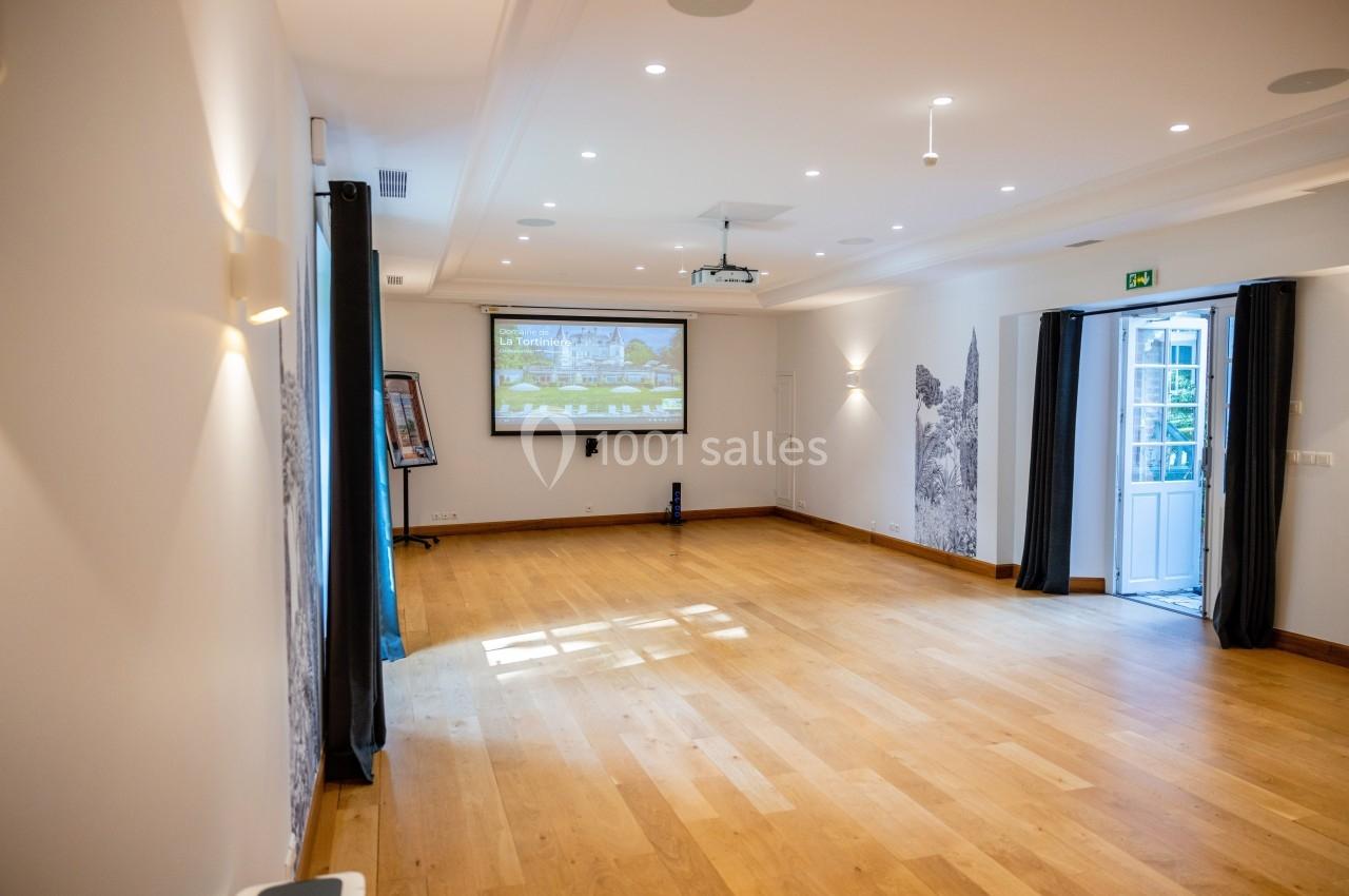 Salle lumineuse avec parquet en bois, écran de projection, murs décorés et porte-fenêtre ouverte sur l'extérieur.