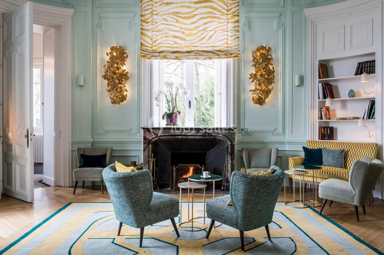 Salon élégant avec cheminée allumée, fauteuils bleus, table basse ronde et décorations dorées sur murs pastel.