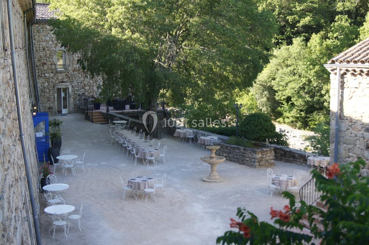 Cour extérieure avec tables et chaises en fer forgé, fontaine centrale, arbres et bâtiments en pierre environnants.