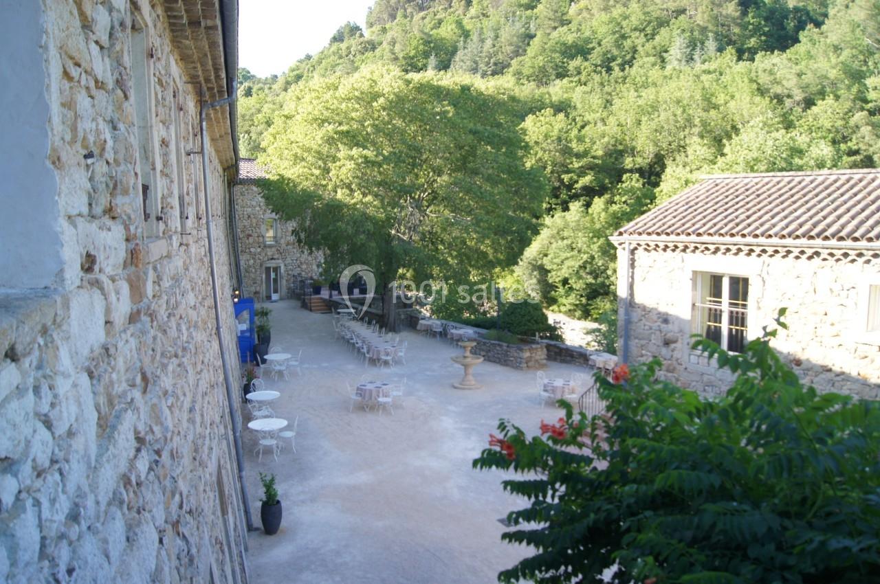 Cour extérieure d'un bâtiment en pierre avec des tables et chaises, entourée de végétation et d'arbres.