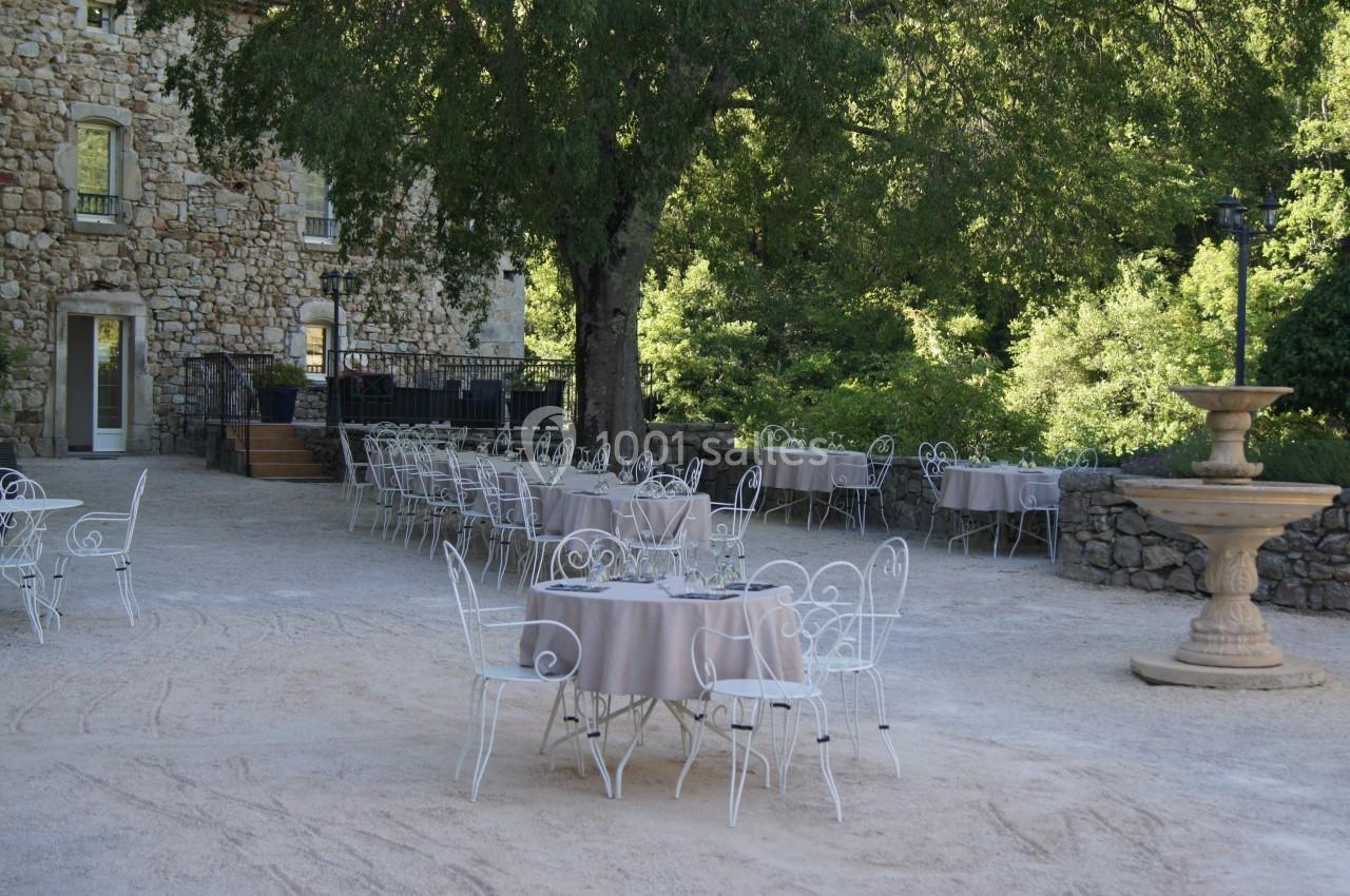 Terrasse extérieure avec tables et chaises en fer forgé, entourée de verdure et d'un bâtiment en pierre.