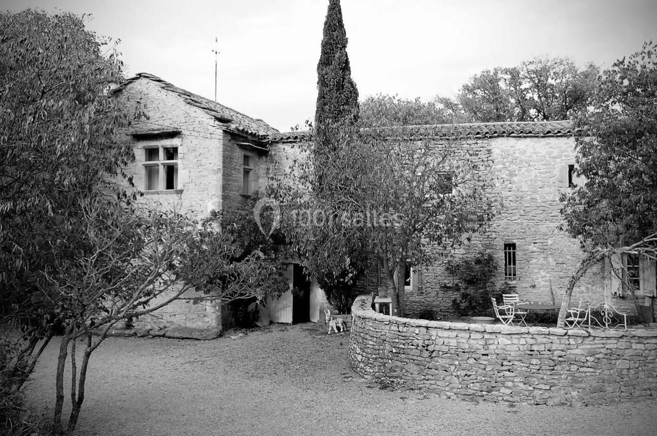 Maison en pierre ancienne entourée d'arbres, avec une cour et un muret en pierre au premier plan.