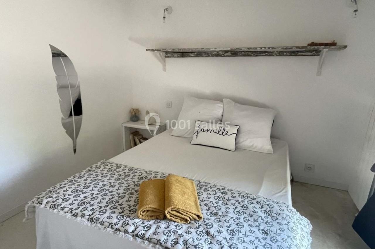 Lit double avec coussins blancs, plaid à motifs et serviettes jaunes pliées, dans une chambre minimaliste avec étagère…