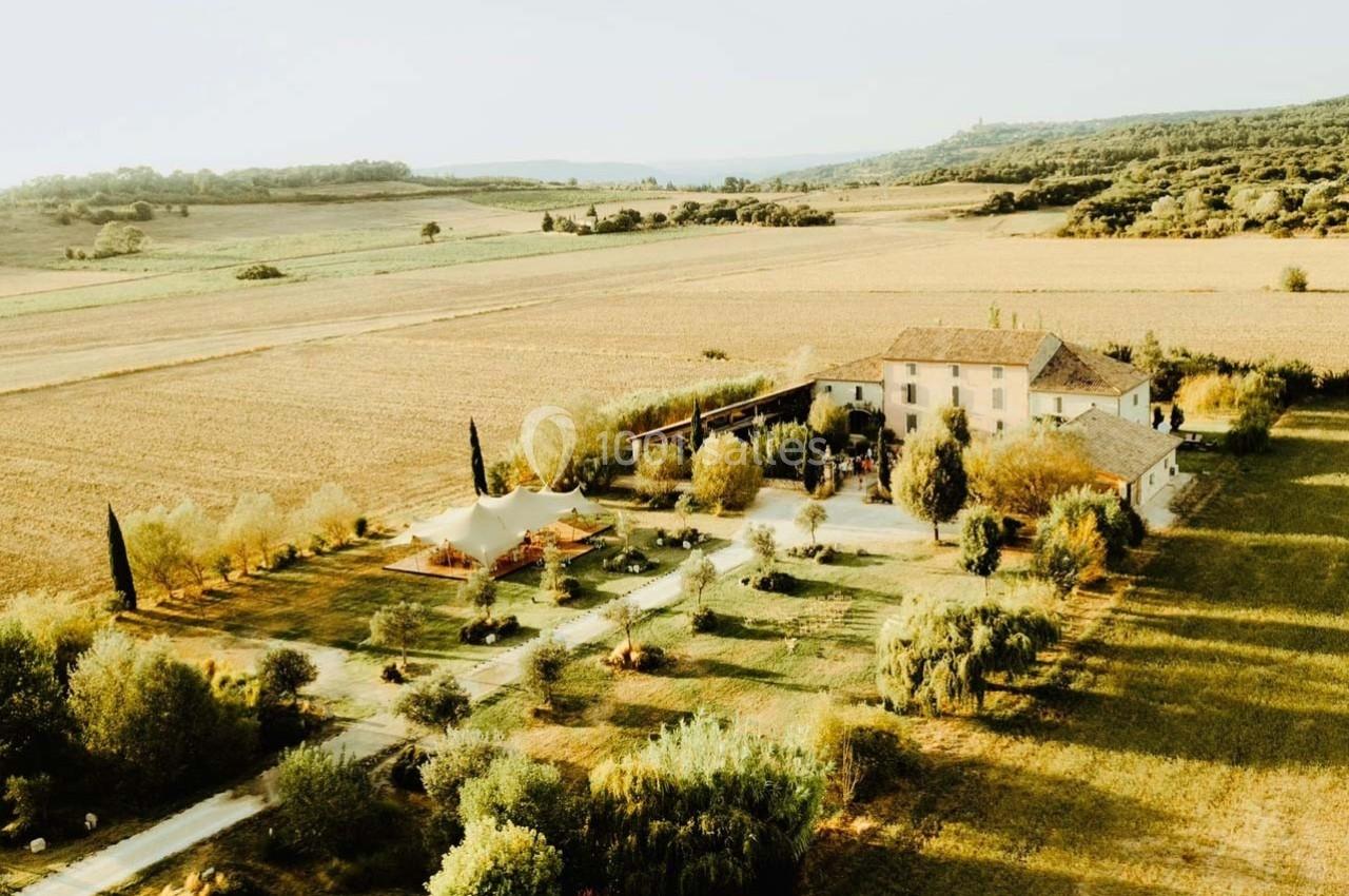 Vue aérienne d'une grande maison entourée de champs et d'arbres dans un paysage rural ensoleillé.