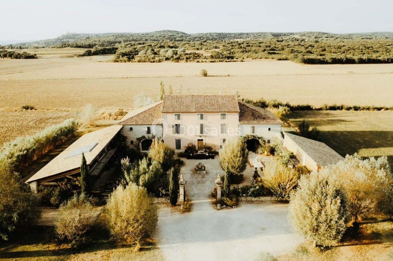 Vue aérienne d'une grande maison en pierre entourée de champs, d'arbres et de bâtiments annexes sous un ciel clair.