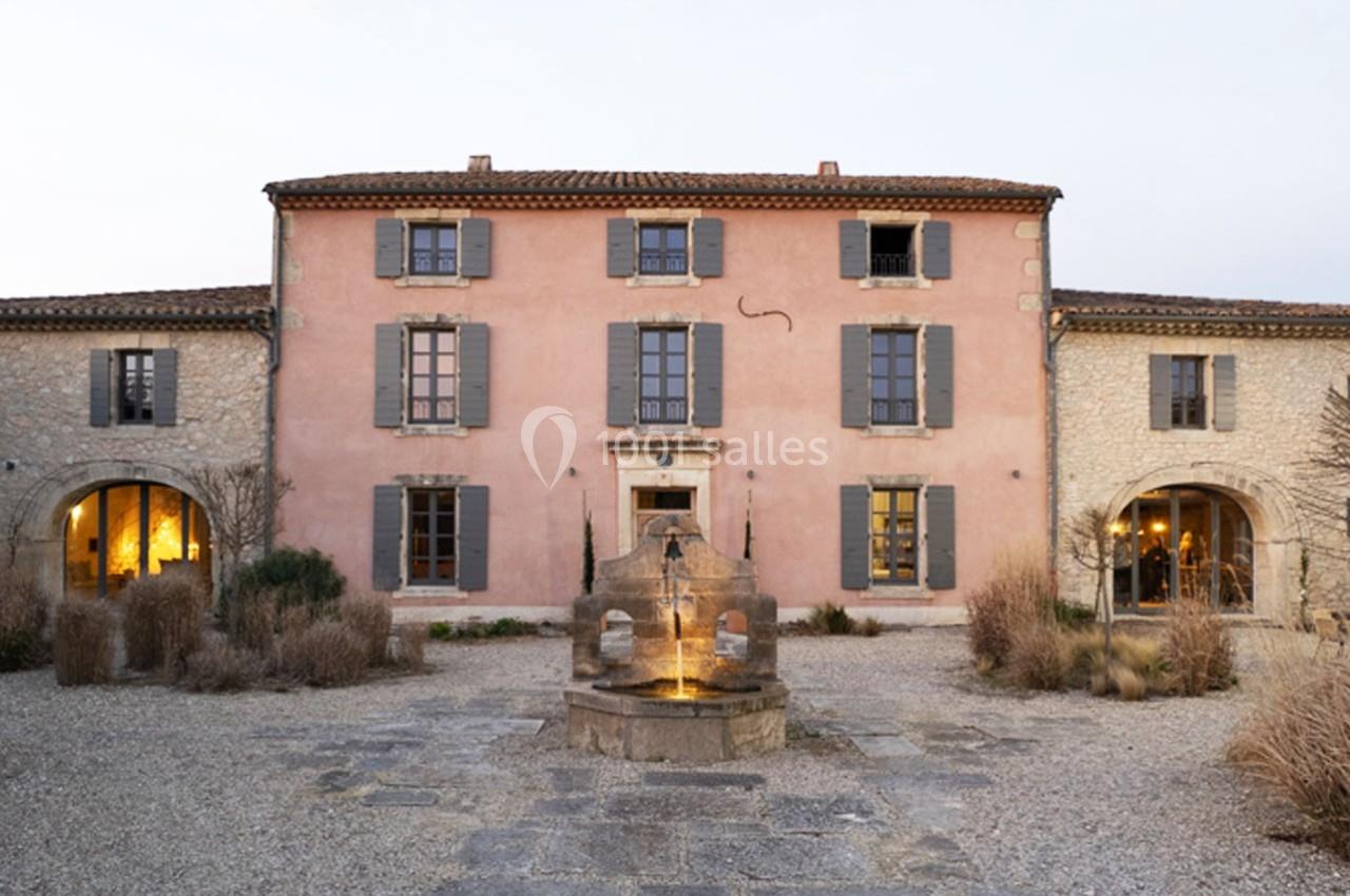 Façade d'une grande maison en pierre avec volets gris, une fontaine centrale et une cour gravillonnée.
