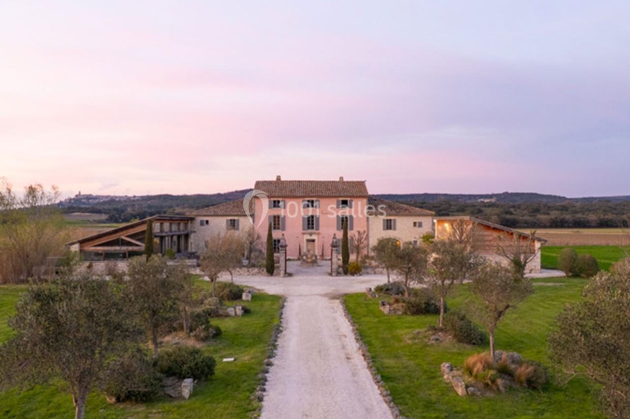 Allée bordée d'oliviers menant à une grande maison en pierre entourée de champs sous un ciel au coucher du soleil.