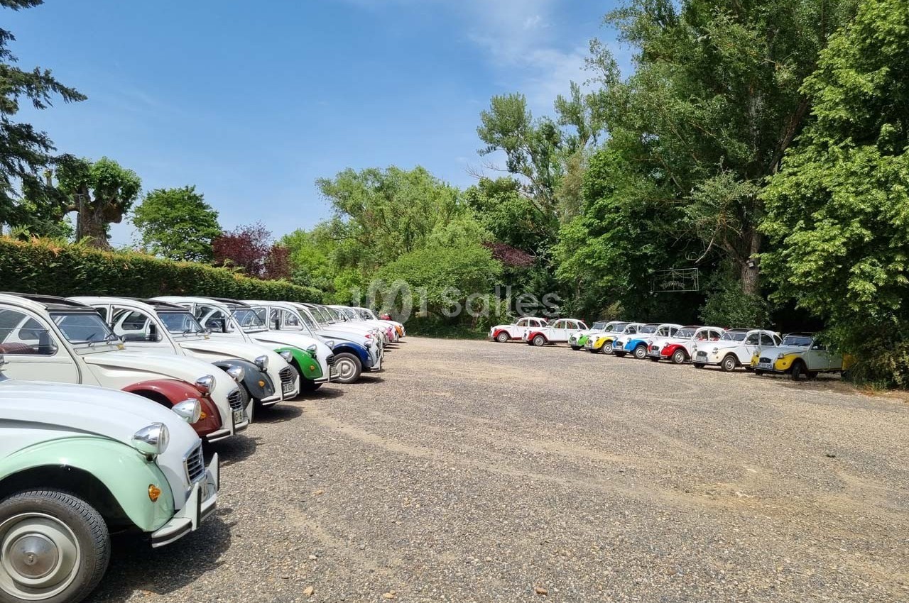 Rangée de voitures anciennes colorées stationnées sur un parking en gravier, entouré de verdure et d'arbres.