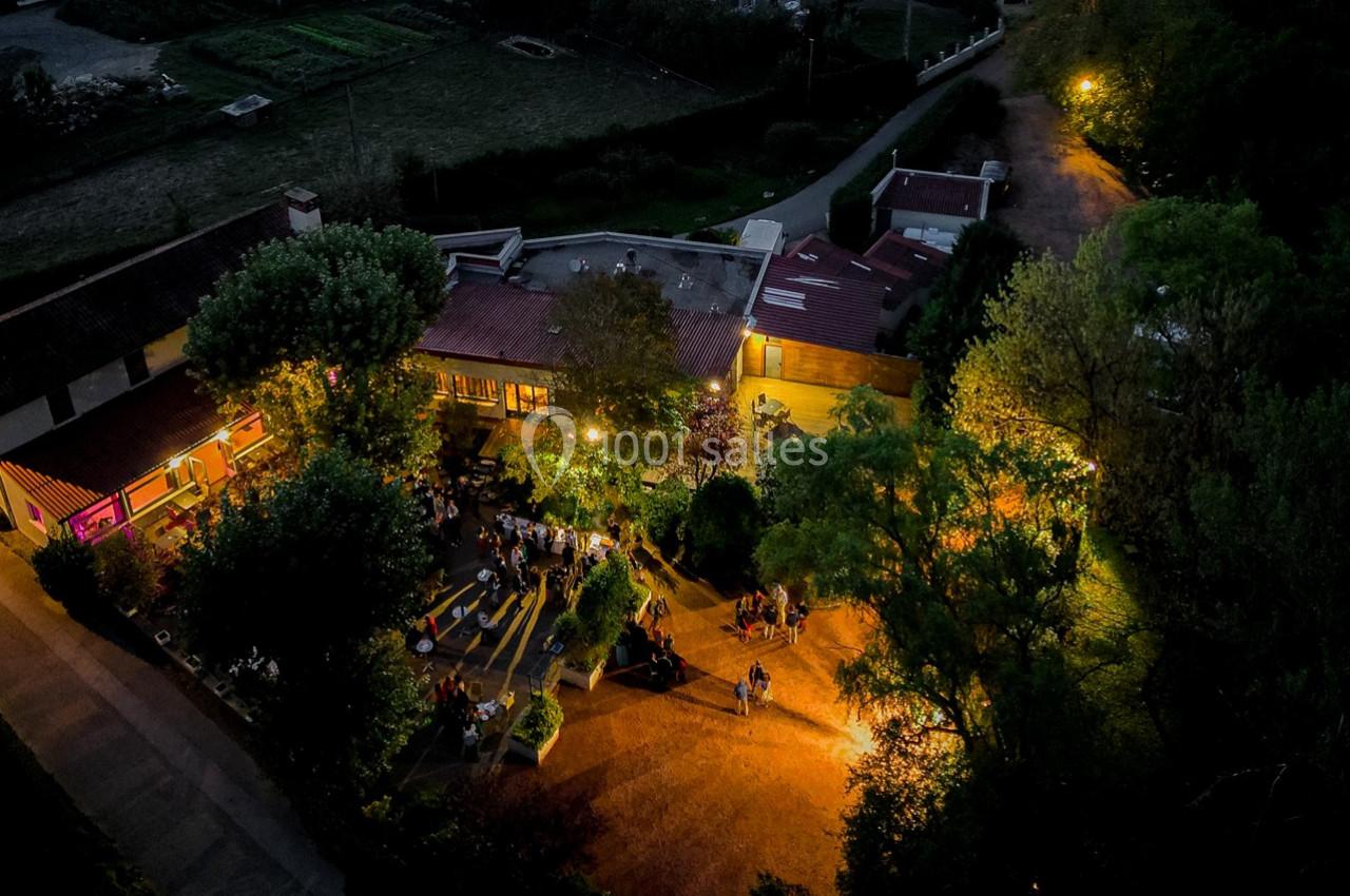 Vue aérienne nocturne d'un espace extérieur éclairé avec des arbres, des bâtiments et des personnes réunies.