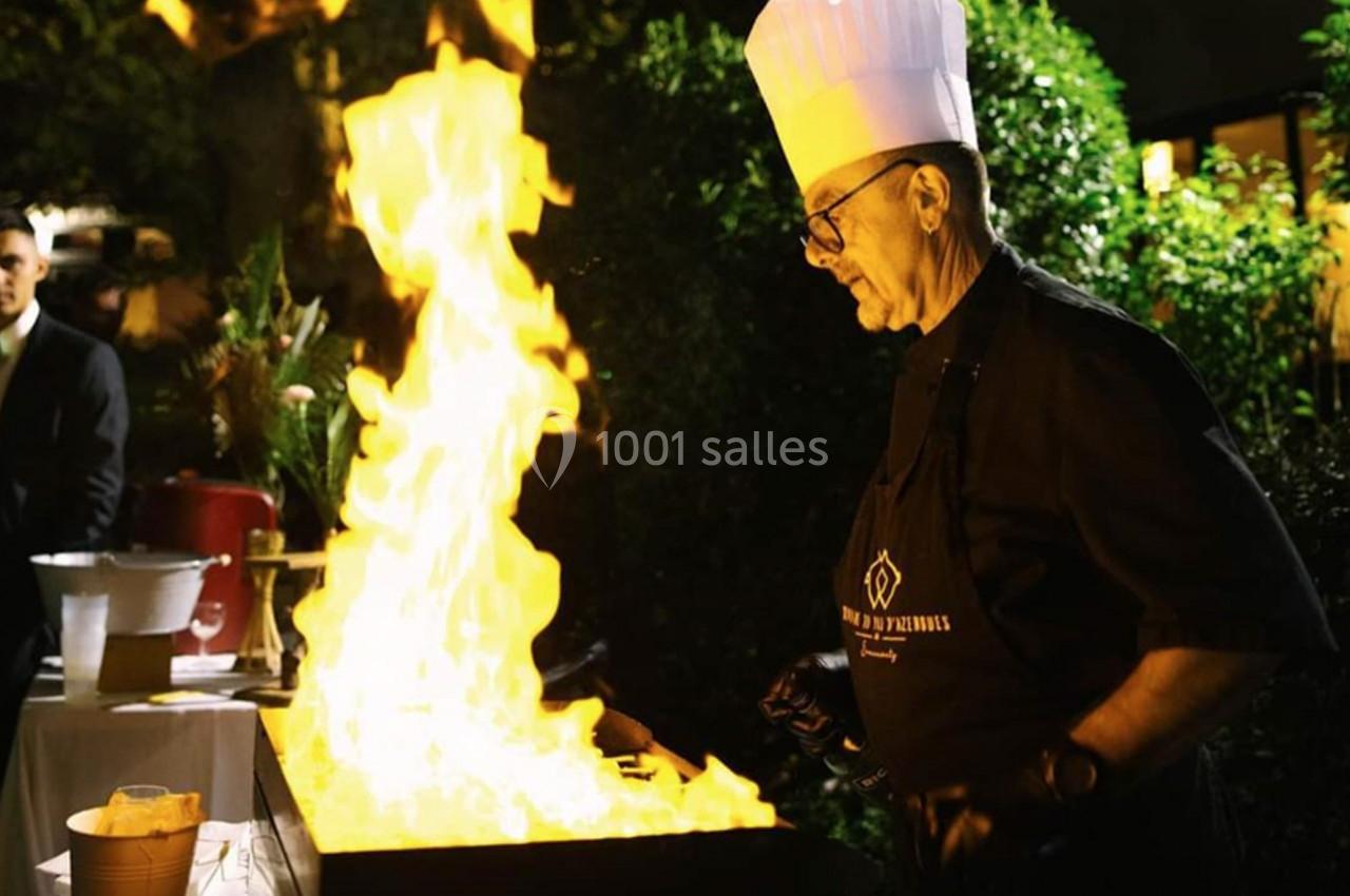 Un chef en toque blanche cuisine devant une grande flamme sur une plaque chauffante en extérieur, de nuit.