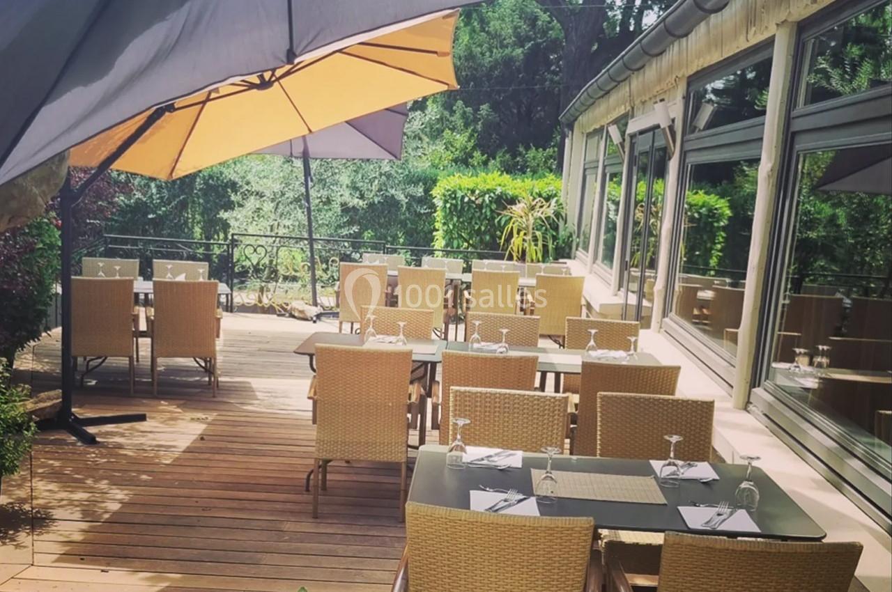 Terrasse en bois avec des tables et chaises en rotin, partiellement ombragée par un grand parasol, entourée de verdure.