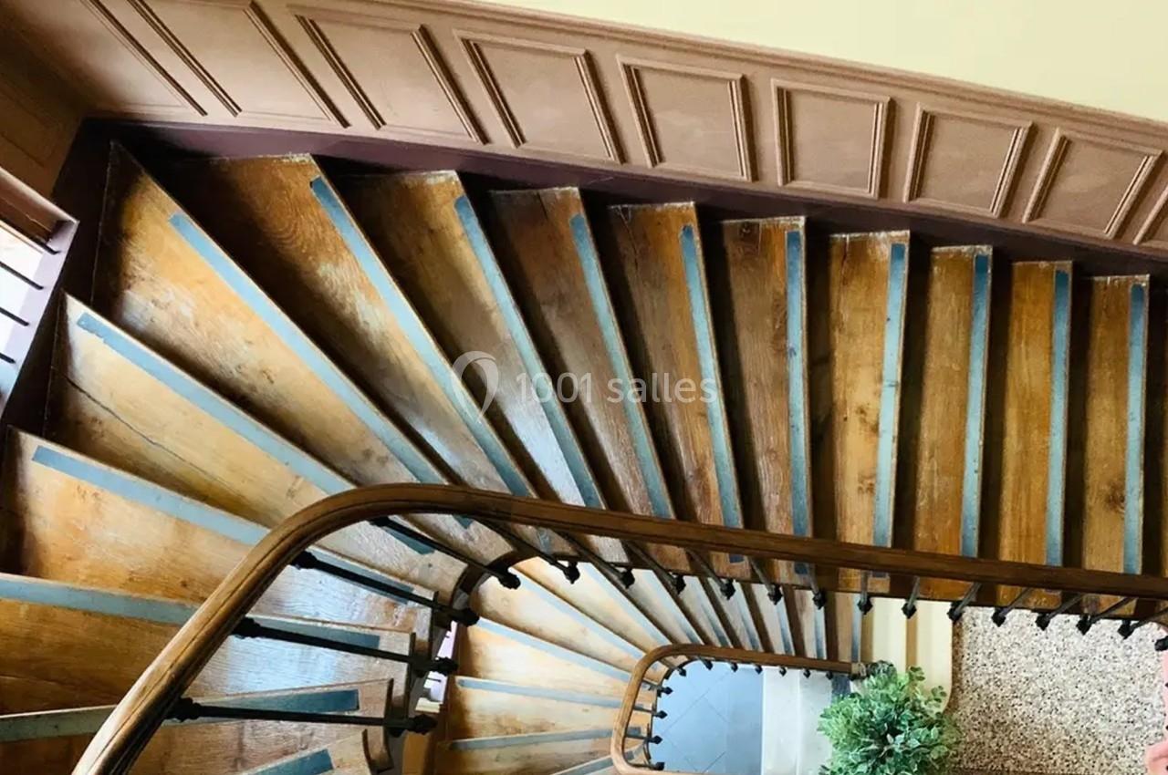 Escalier en bois vu de haut, avec une rampe courbée et des marches usées, entouré de murs clairs.