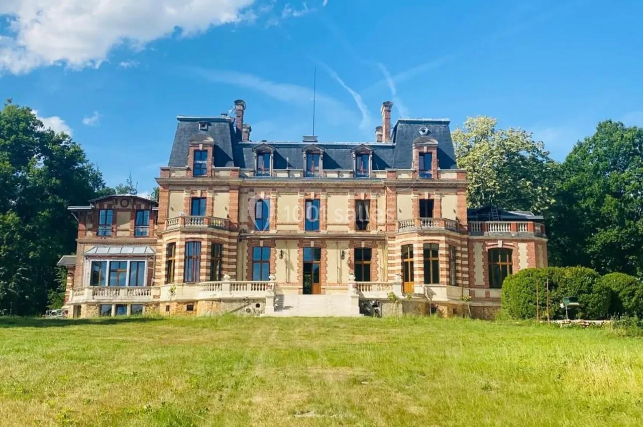 Façade d'un château en briques rouges avec toit mansardé, entouré de pelouse et d'arbres sous un ciel bleu.