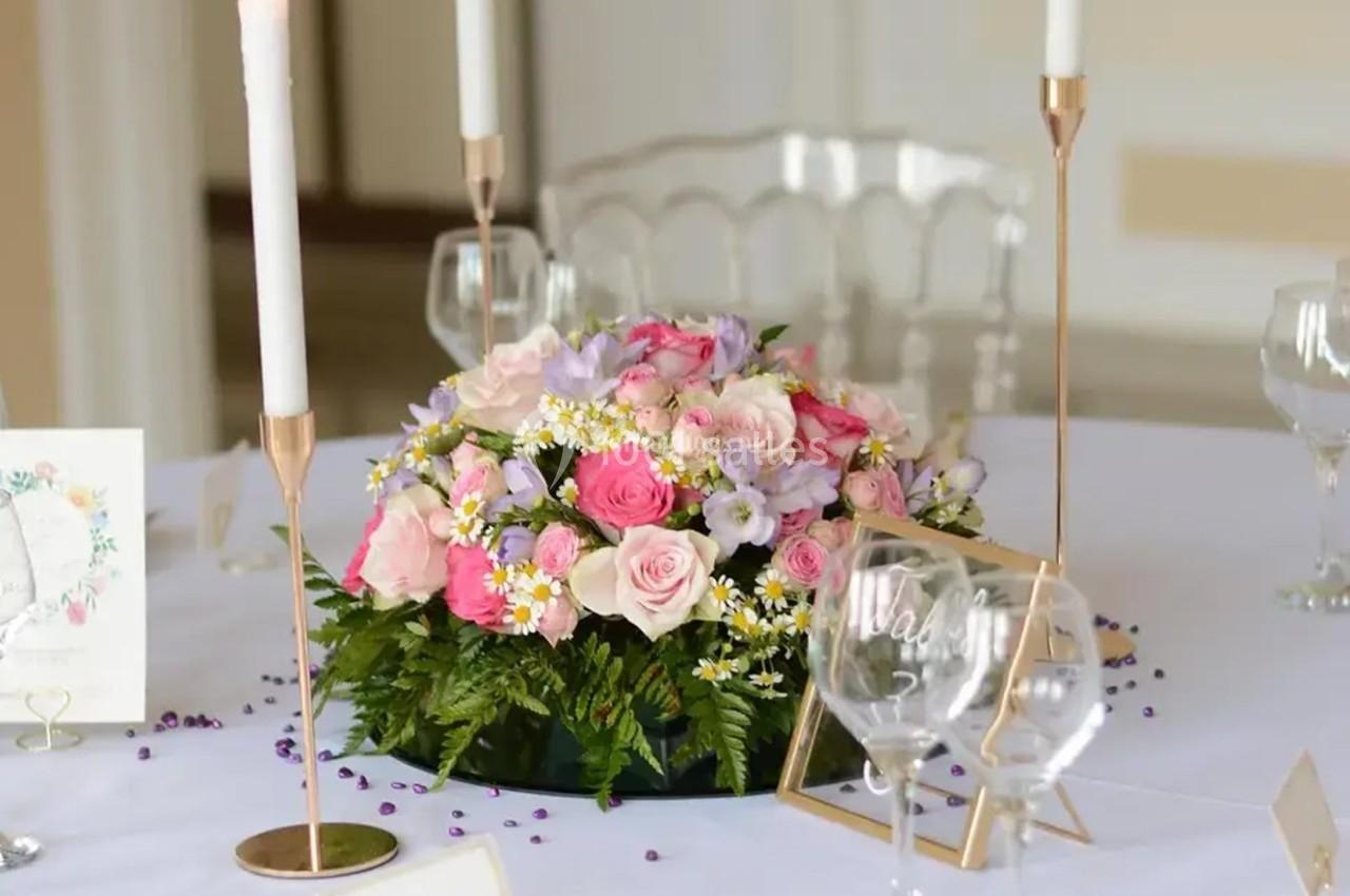 Centre de table floral avec roses roses, fleurs violettes et blanches, entouré de bougies et de verres sur une nappe blanche.