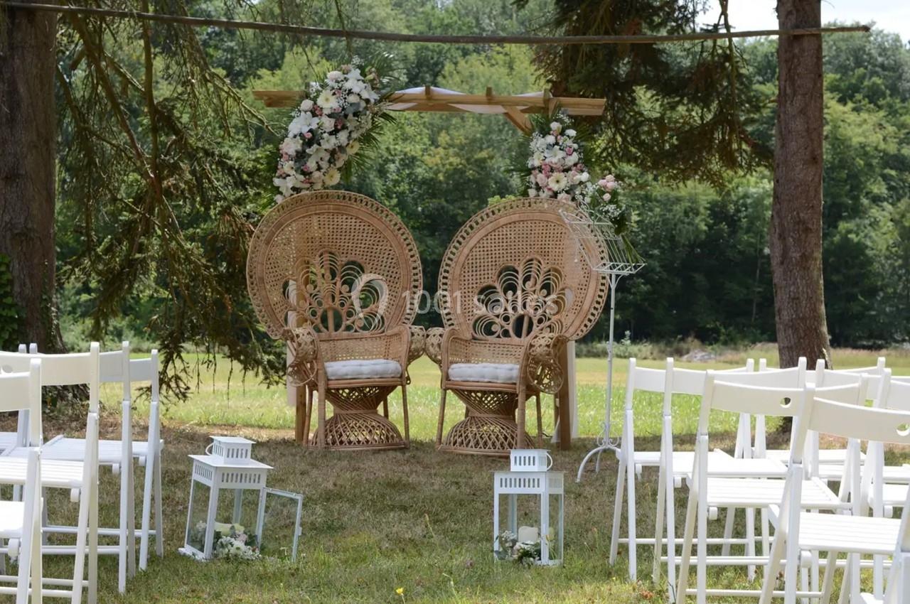 Deux fauteuils en rotin ornés de fleurs sous une arche décorée, entourés de chaises blanches dans un cadre extérieur…