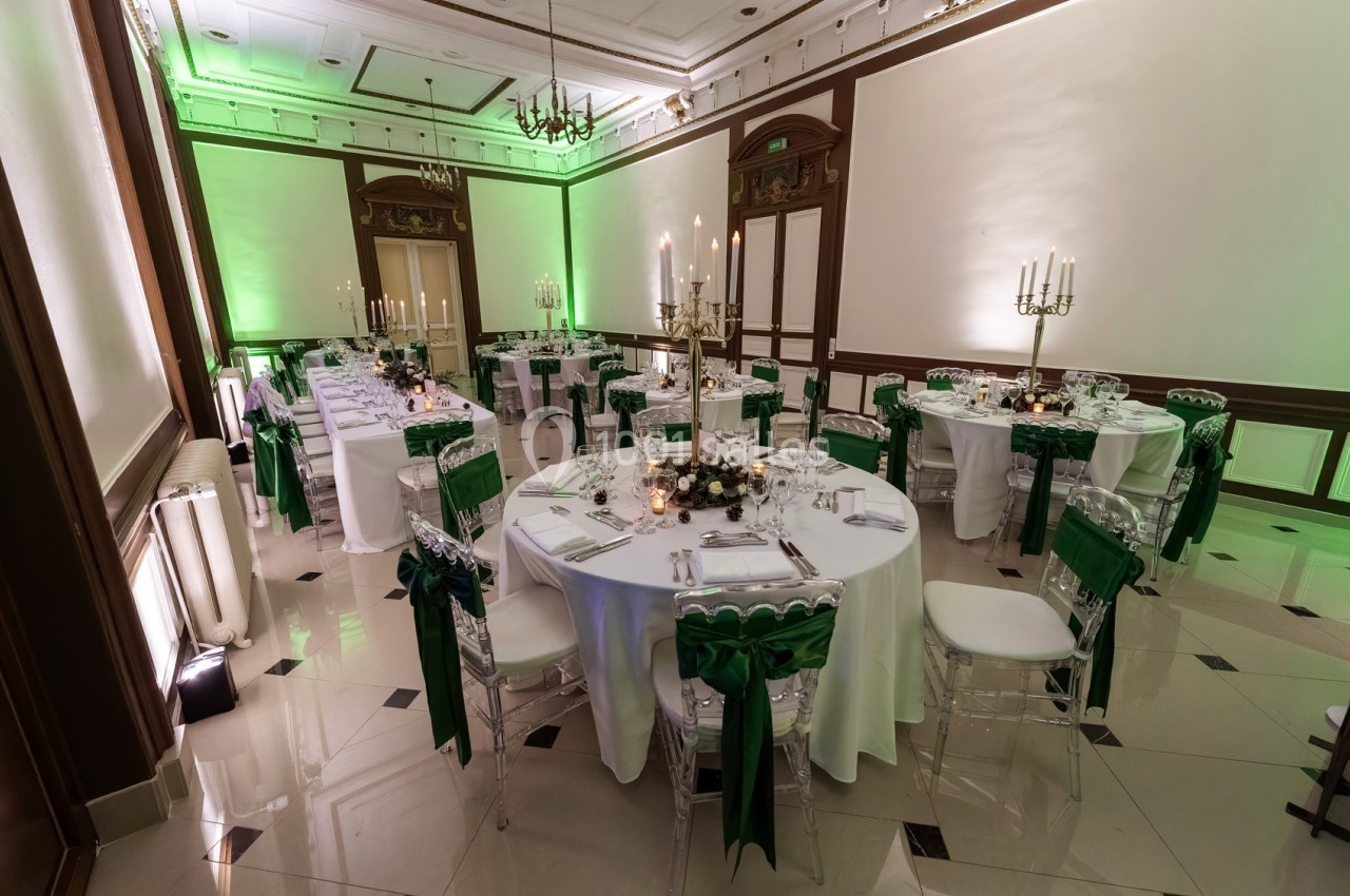 Salle de réception élégante avec tables rondes dressées, chaises transparentes ornées de nœuds verts et éclairage tamisé.