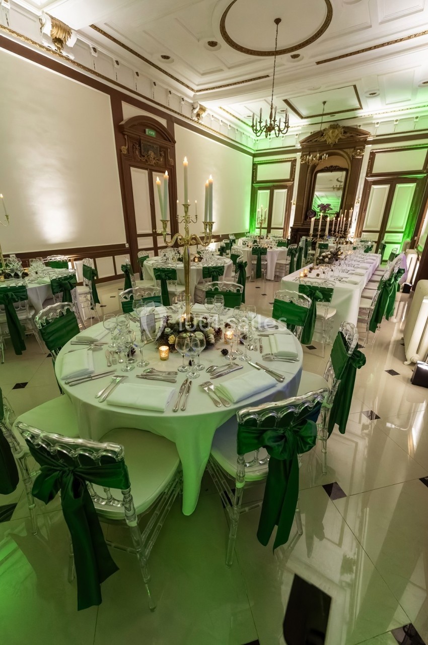 Salle de réception élégante avec tables rondes dressées, chaises transparentes ornées de nœuds verts et éclairage chaleureux.