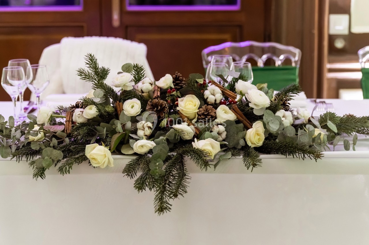 Centre de table floral avec roses blanches, branches de sapin, pommes de pin et coton, posé sur une nappe blanche.
