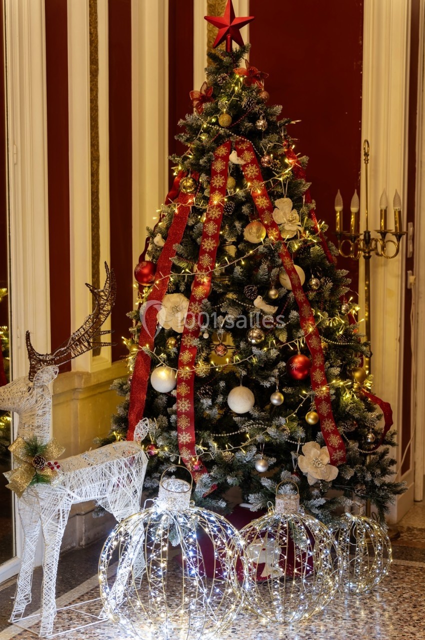 Sapin de Noël décoré de guirlandes rouges, boules dorées et blanches, entouré de décorations lumineuses et rennes blancs.