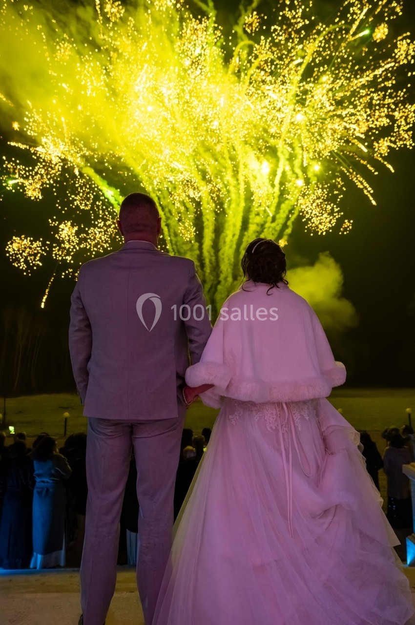 Un couple en tenue de mariage regarde un feu d'artifice jaune éclatant dans le ciel nocturne.