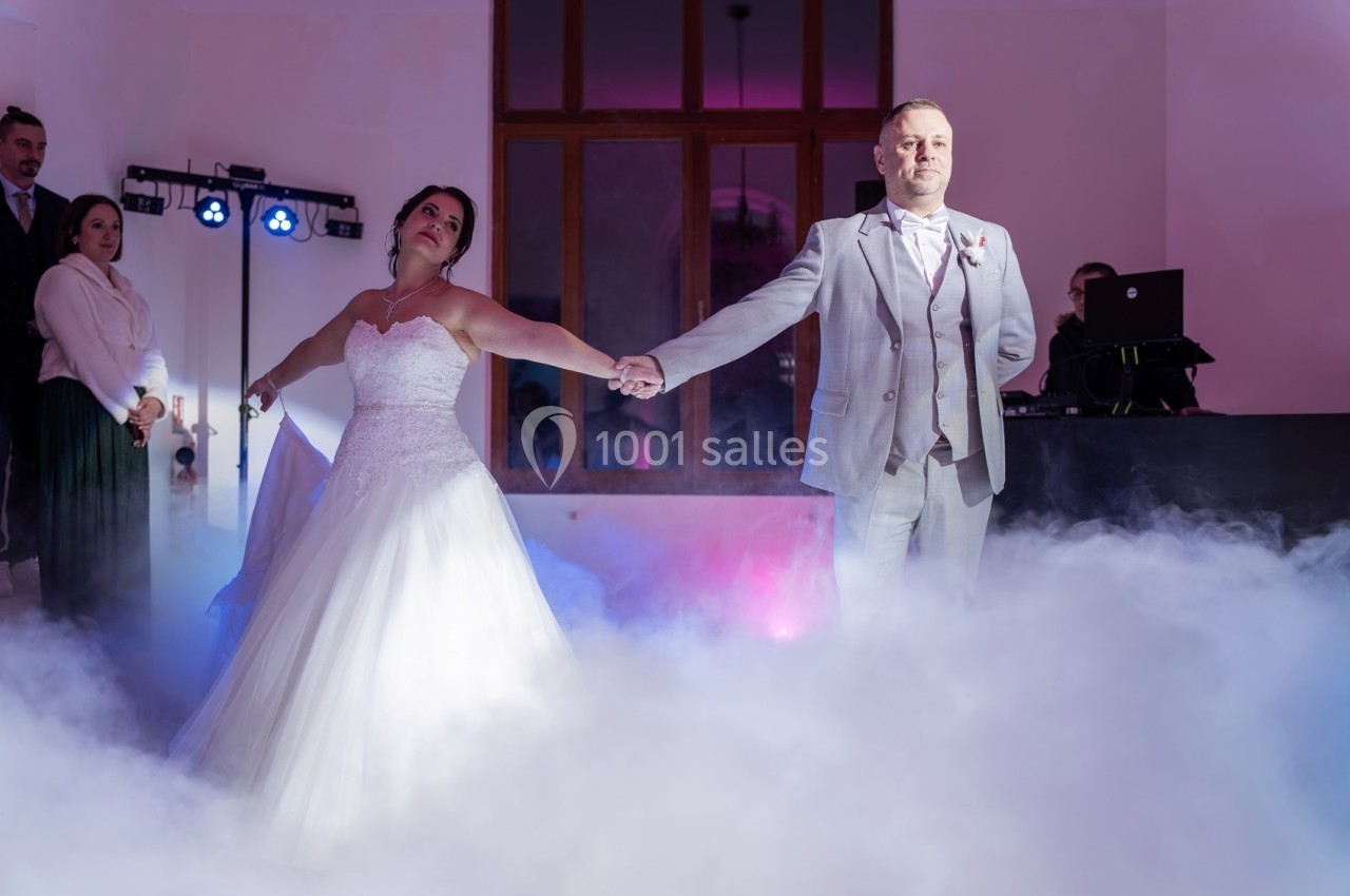 Un couple en tenue de mariage danse sur une piste entourée de fumée, éclairée par des lumières colorées.