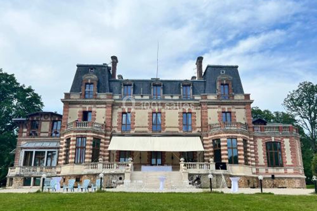 Façade d'un grand bâtiment historique en briques rouges et pierres, entouré de verdure sous un ciel partiellement nuageux.