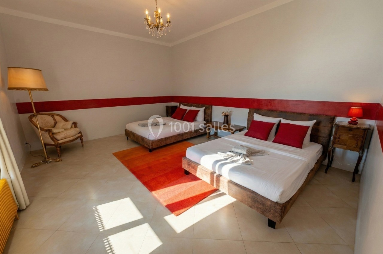 Chambre lumineuse avec deux lits doubles, décoration simple, tapis orange, lampe et fauteuil en osier.