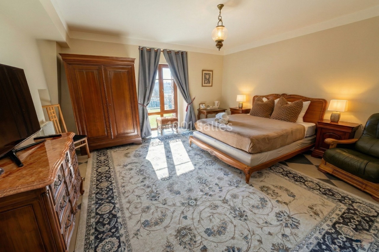 Chambre lumineuse avec grand lit, armoire en bois, fauteuil en cuir, tapis décoratif et fenêtre donnant sur l'extérieur.
