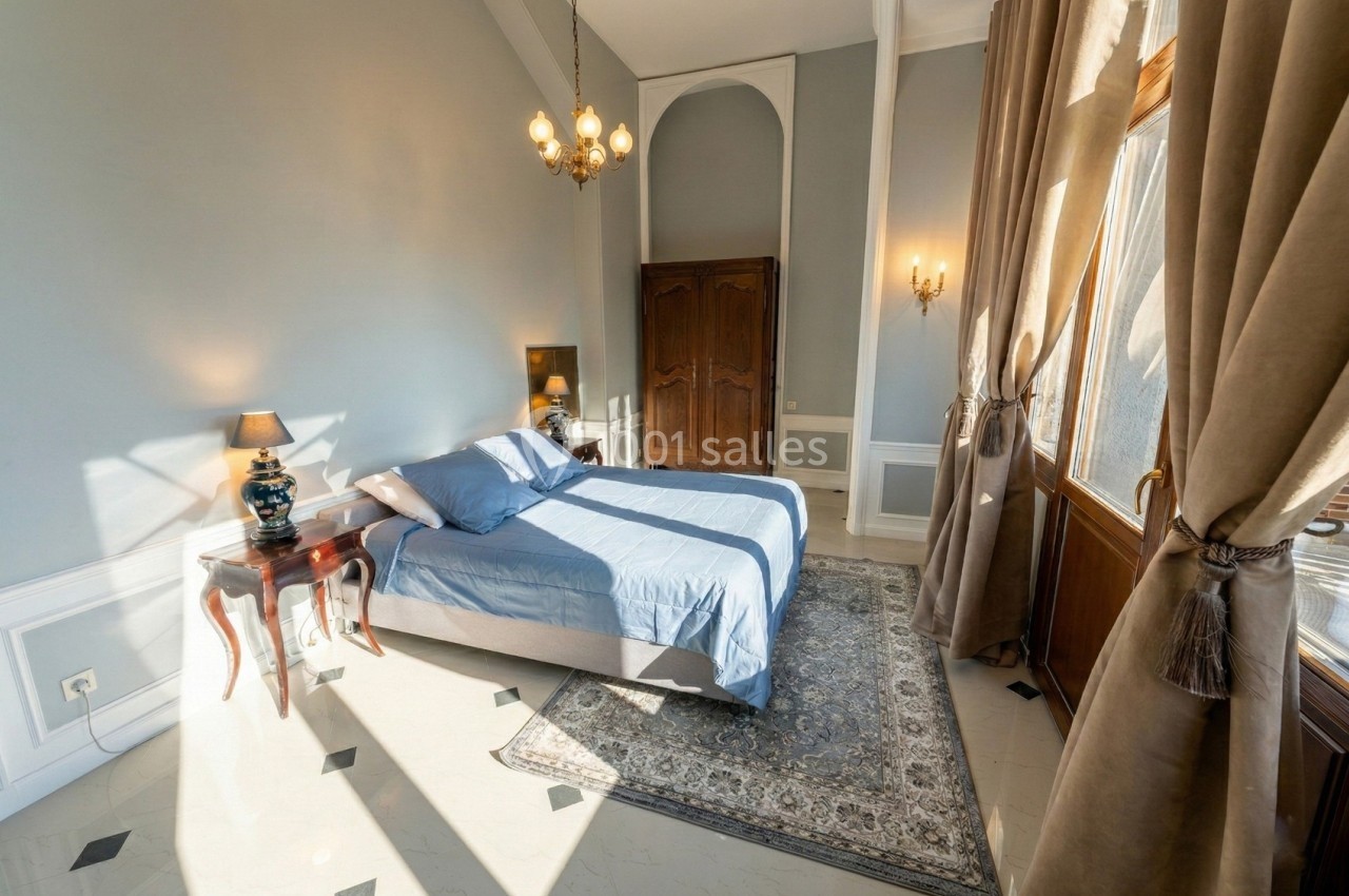Chambre lumineuse avec lit double, draps bleus, meubles en bois, tapis décoratif et grandes fenêtres avec rideaux beiges.