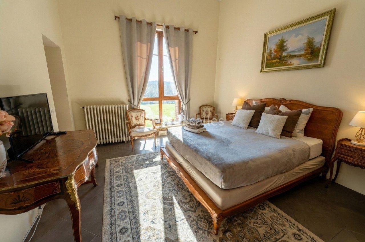 Chambre lumineuse avec lit double, meubles en bois sculpté, tapis décoratif et grande fenêtre avec rideaux.