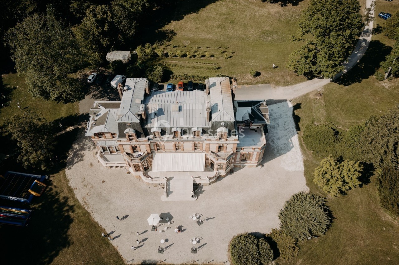 Vue aérienne d'un manoir entouré de jardins, d'arbres et d'une allée gravillonnée, avec quelques personnes au premier plan.