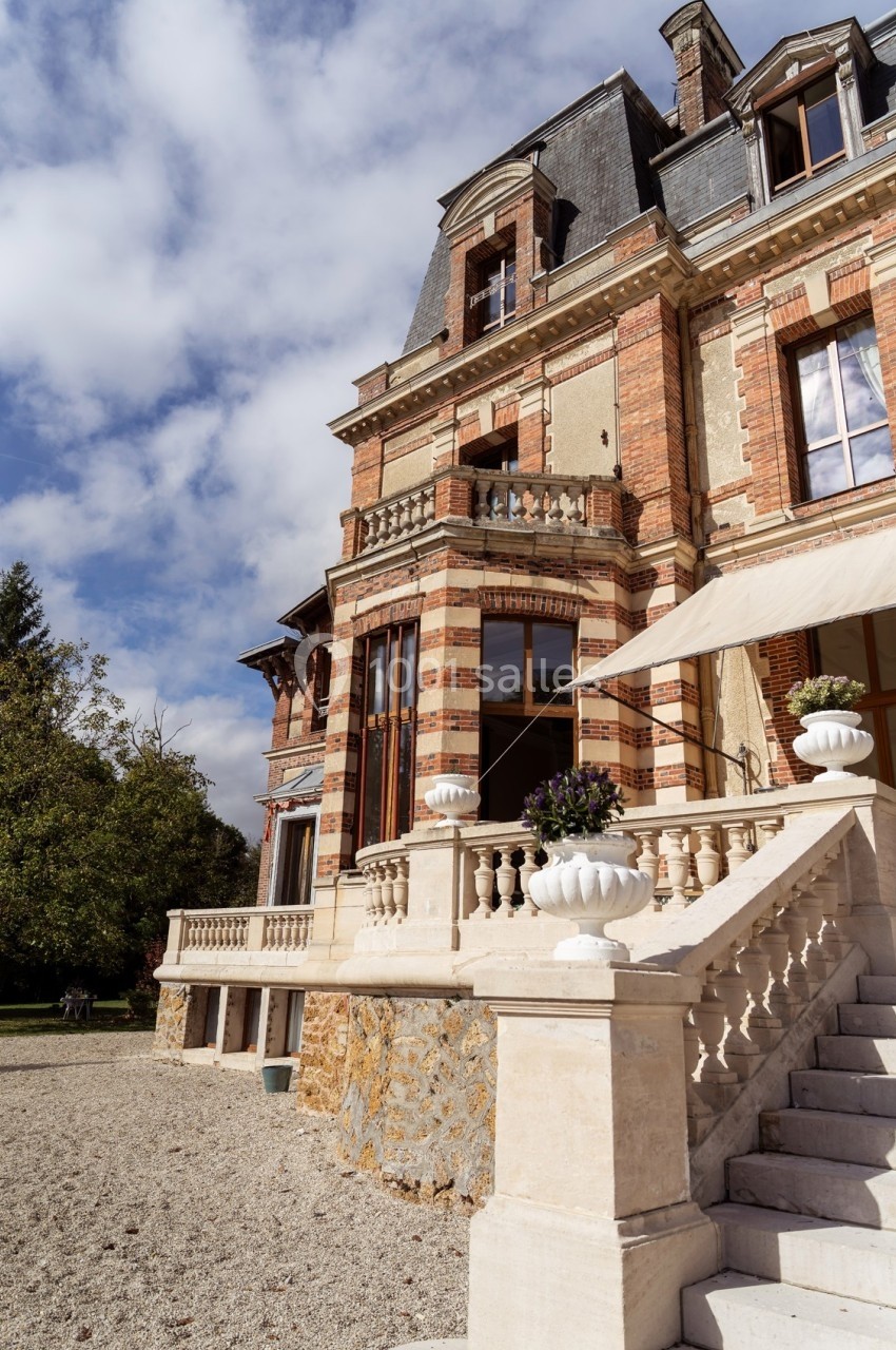 Façade d'un manoir en briques avec escaliers en pierre, balustrade et jardinières, sous un ciel partiellement nuageux.