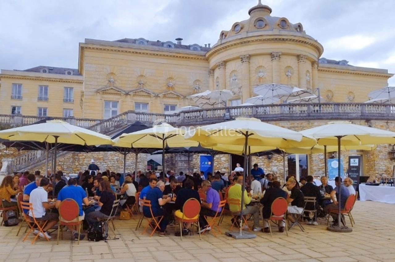 Groupe de personnes assises sous des parasols dans une cour pavée devant un bâtiment historique.