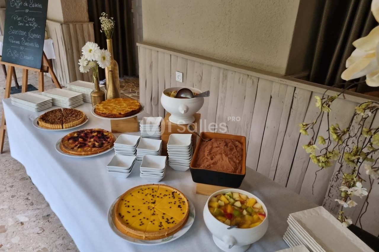 Buffet de desserts avec tartes, mousse au chocolat, salade de fruits et piles d'assiettes sur une table blanche.