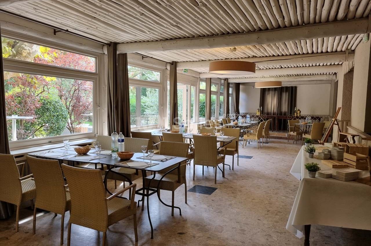 Salle lumineuse avec grandes baies vitrées, tables dressées et buffet installé, entourée de verdure extérieure.