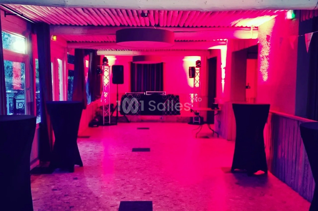 Salle de réception éclairée par des lumières rouges, avec tables hautes et espace pour un DJ au fond.