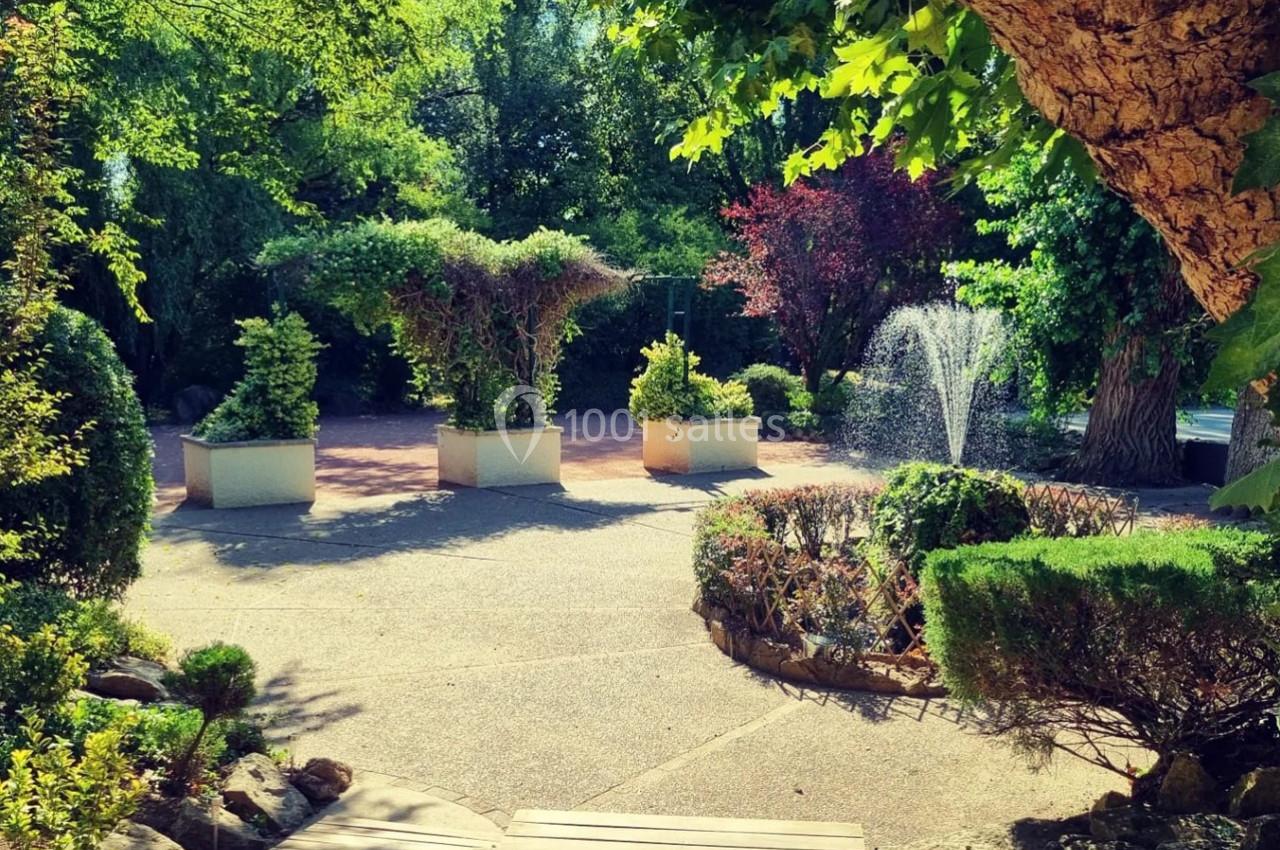 Jardin avec allée pavée, fontaine en arrière-plan et arbustes taillés dans des bacs sous un ciel ensoleillé.