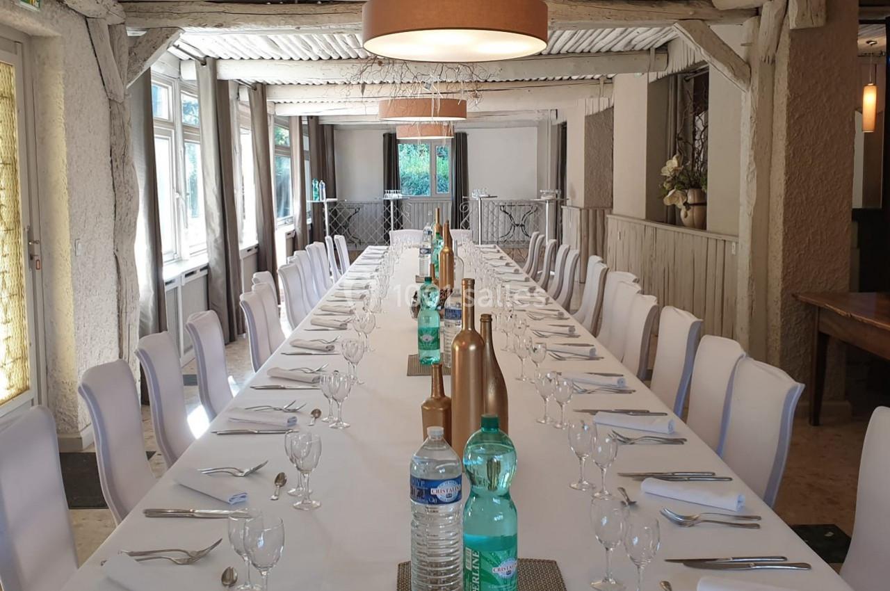 Grande table rectangulaire dressée pour un repas, entourée de chaises blanches, dans une salle lumineuse au style rustique.