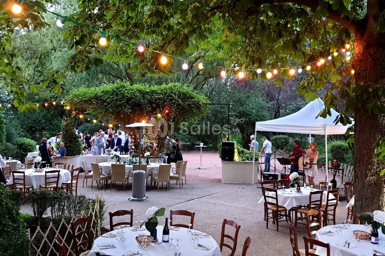 Terrasse extérieure avec tables dressées, guirlandes lumineuses, musiciens et invités dans un cadre verdoyant.
