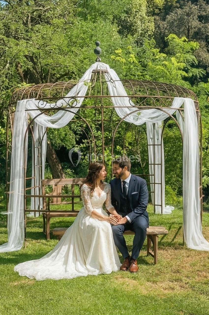 Un couple assis sous une tonnelle décorée de voilages blancs dans un jardin verdoyant.