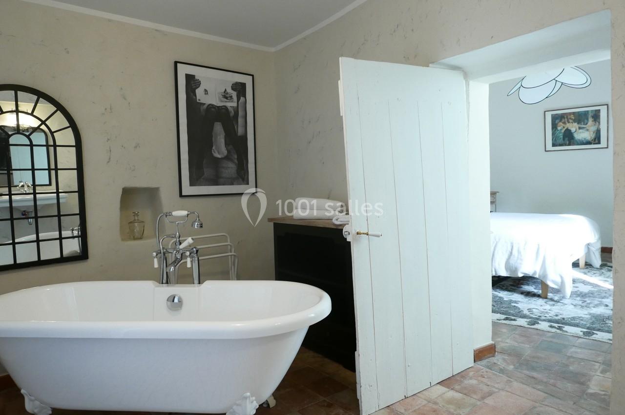 Salle de bain avec baignoire sur pieds, miroir en arche, et porte ouverte donnant sur une chambre lumineuse.