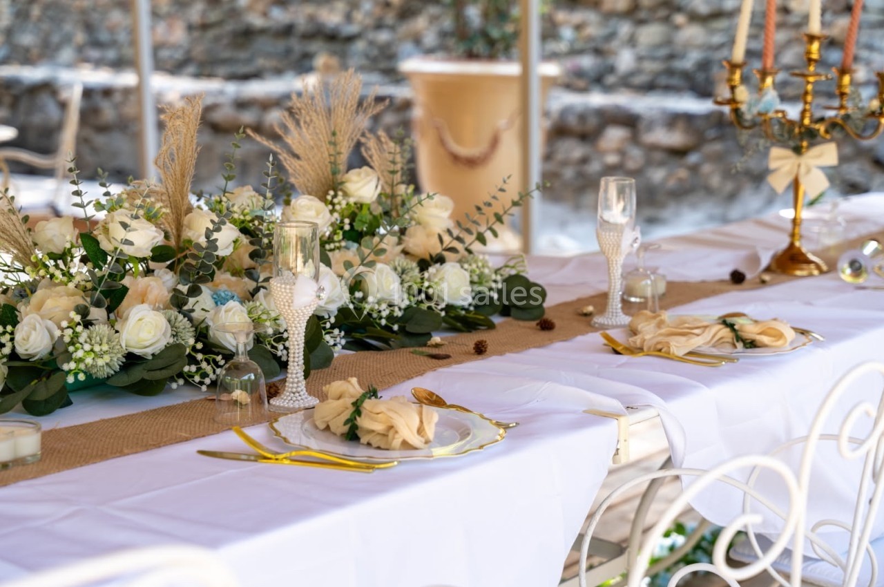 Table décorée pour un événement, avec nappes blanches, fleurs, bougies et couverts dorés dans un cadre extérieur.