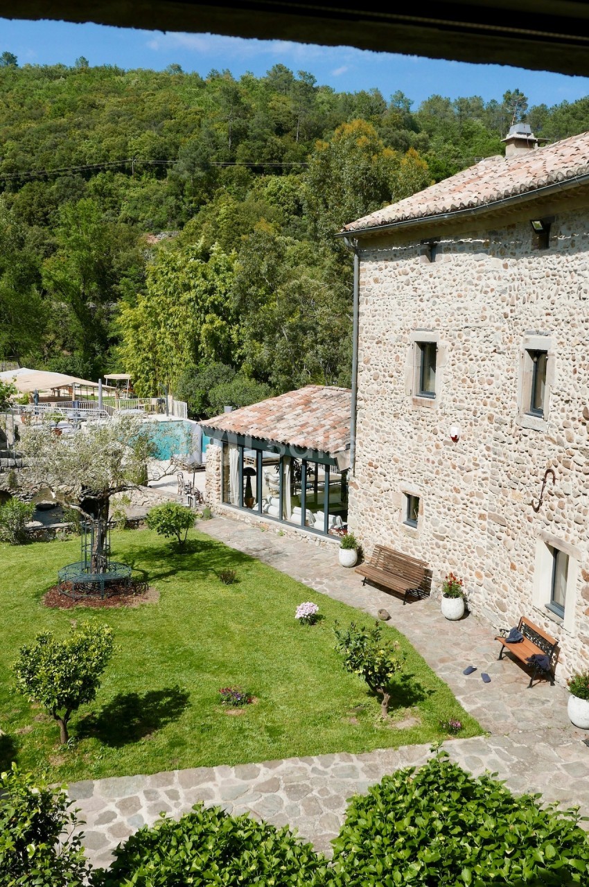 Cour intérieure d'une maison en pierre avec jardin verdoyant, arbres fruitiers, bancs et piscine en arrière-plan.