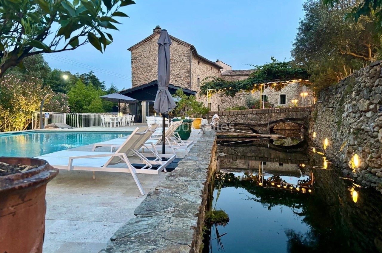 Piscine extérieure avec transats, maison en pierre et végétation environnante reflétée dans un bassin au crépuscule.
