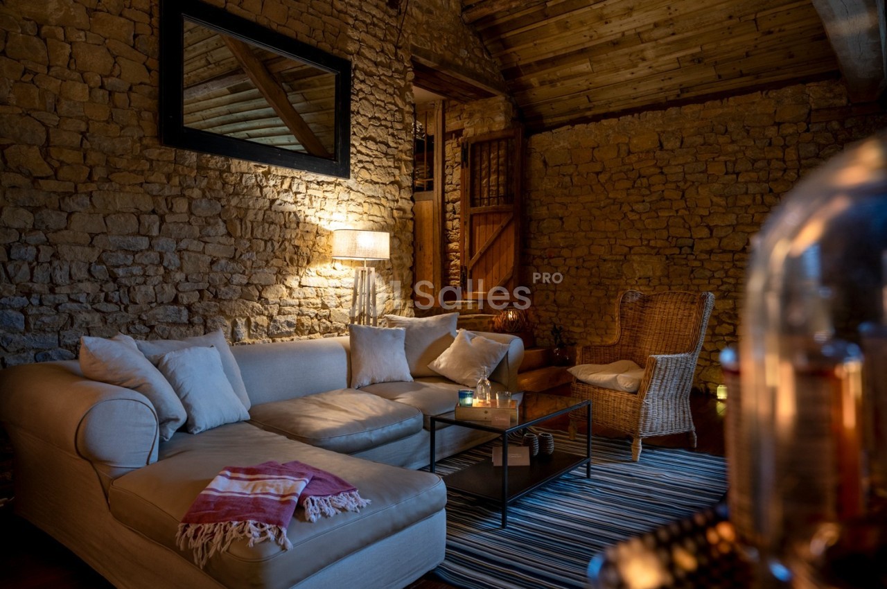 Salon chaleureux avec murs en pierre, canapé beige, fauteuil en osier, éclairage tamisé et décoration cosy.