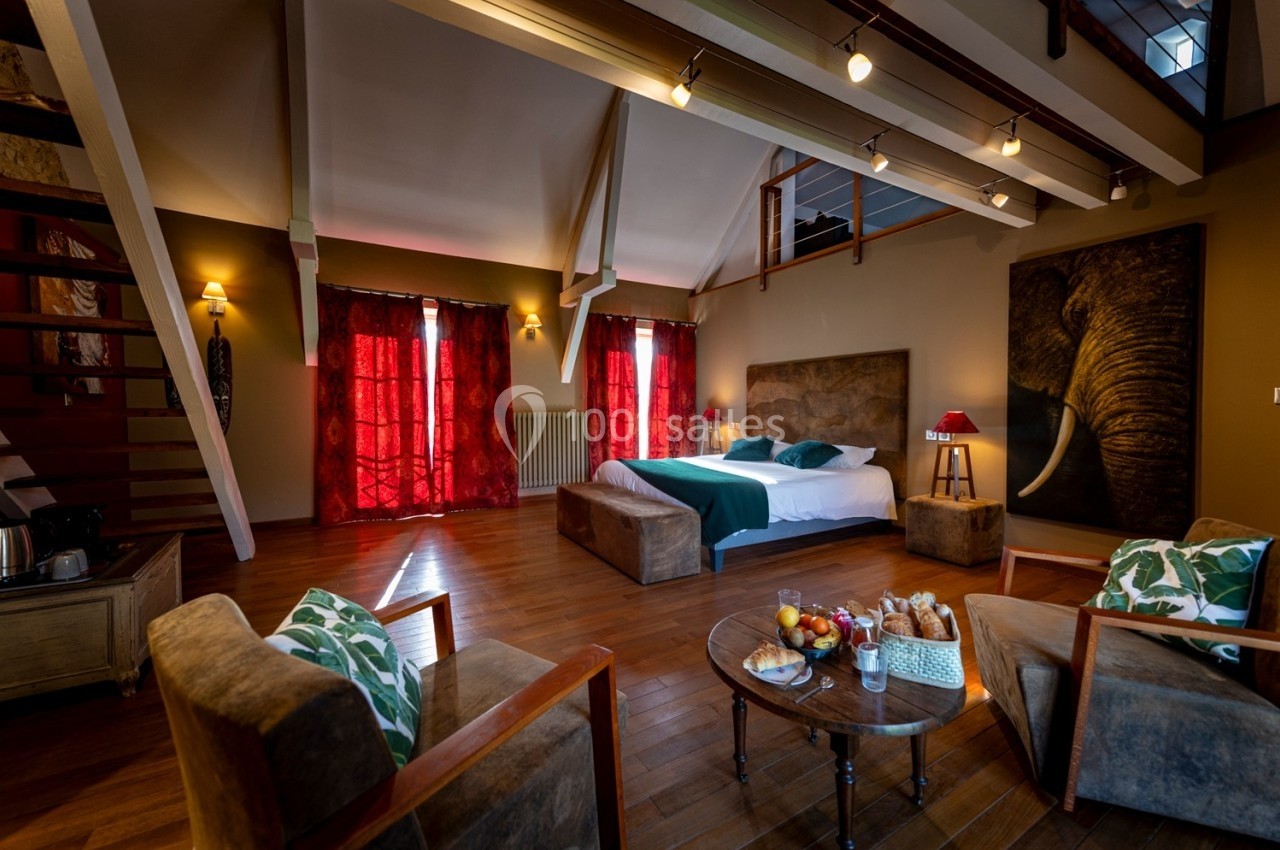 Chambre spacieuse avec lit double, fauteuils, table basse garnie et décor chaleureux sous un plafond en poutres apparentes.