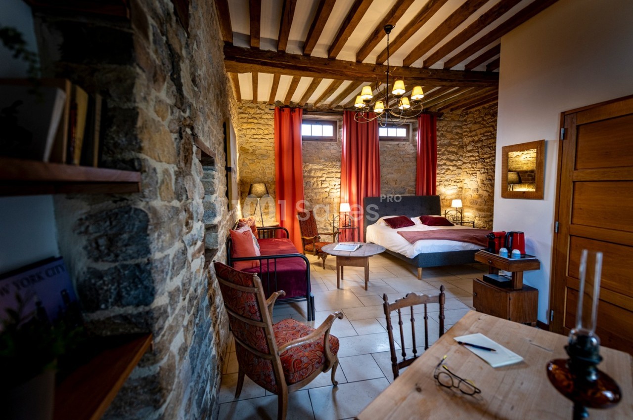 Chambre chaleureuse avec murs en pierre, lit double, fauteuils, bureau en bois et lumière naturelle.